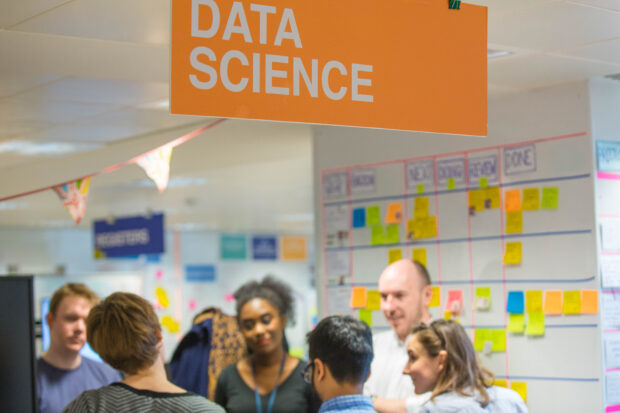 Six people in conversation during a stand-up ceremony in an office, underneath a banner which says 'Data Science'.