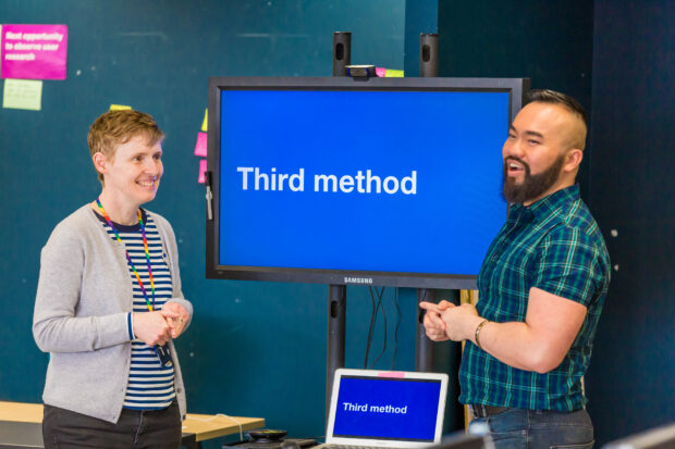 Louise and Haur stand either side of a TV screen, about to present to team colleagues about the 'Third method'