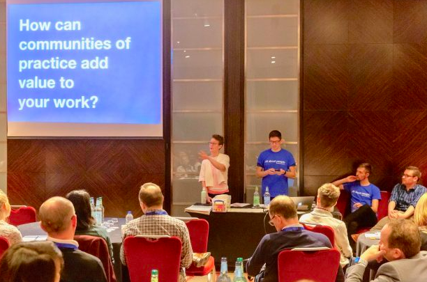 Image of two people presenting at a conference, in front of a screen showing 'How can communities of practice add value to your work'