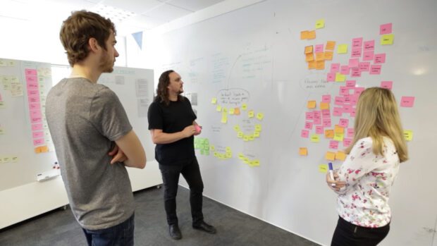 Three people are in an office all staring intently at sticky notes stuck to a giant white board. The sticky notes are dotted around the text "Prevent Fraud and Error".