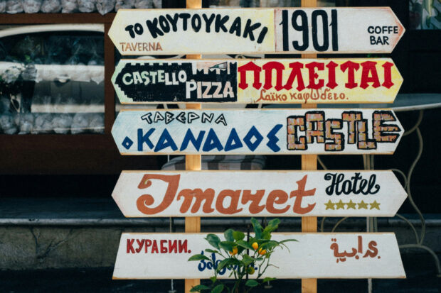 A stack of signposts pointing in different directions bearing words in several different languages. Some of the signposts read: To Koytoykaki Taverna, 1901 Coffee bar, Castello Pizza, Taverna Kanadoe Castle, Imaret Hotel